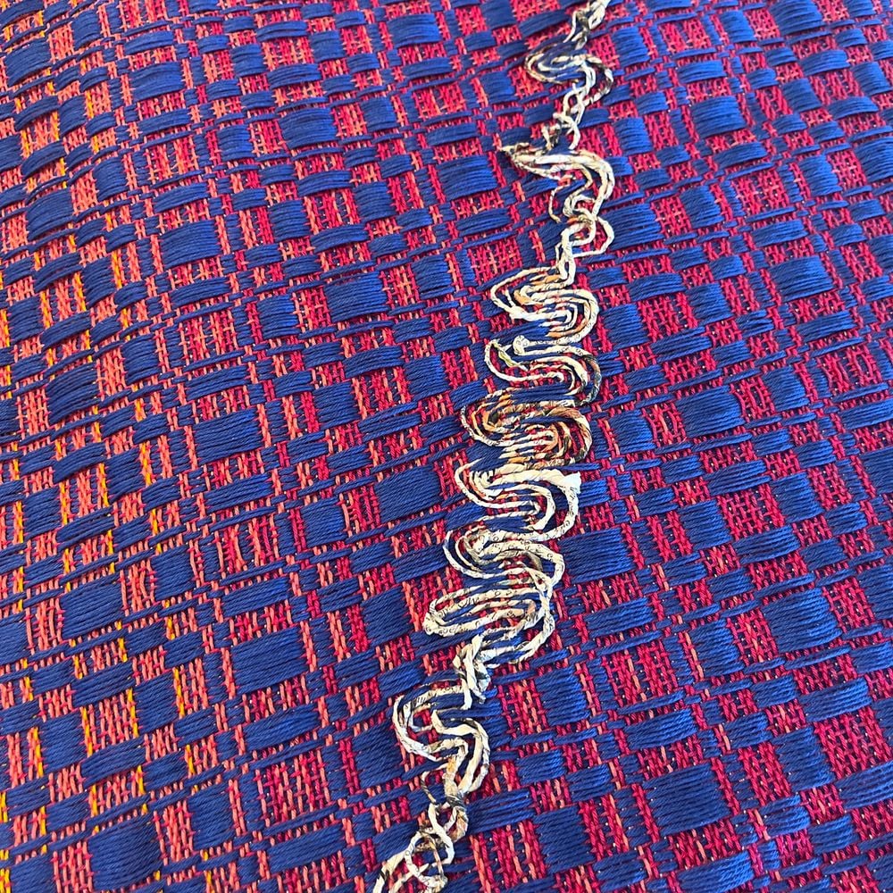 Détail du tissu : un fil de papier journal torsadé à la main serpente en relief à travers l’armure rouge et bleue.